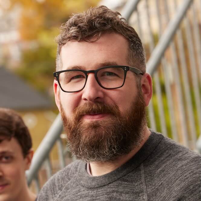 Portrait of Greg Roughton, outdoors, wearing square black glasses and a grey sweater.